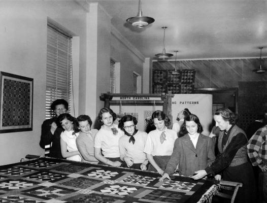 A black and white photo shows seven young people looking at a quilt on a table. An eighth person instructs them.