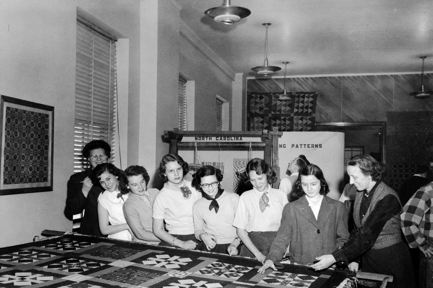 A black and white photo shows seven young people looking at a quilt on a table. An eighth person instructs them.