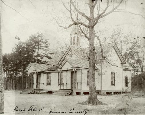 Sepia toned photo of a white schoolhouse. A tree sits in the foreground of the photo.