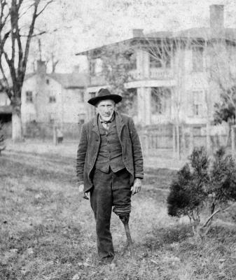 Black and white photo of a man standing in front of a house. His left leg has been amputated and he stands on a peg leg. 