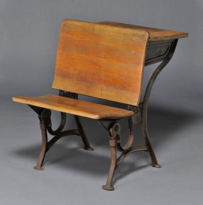 A wooden bench with iron legs and a small desk attached to the back of it. 
