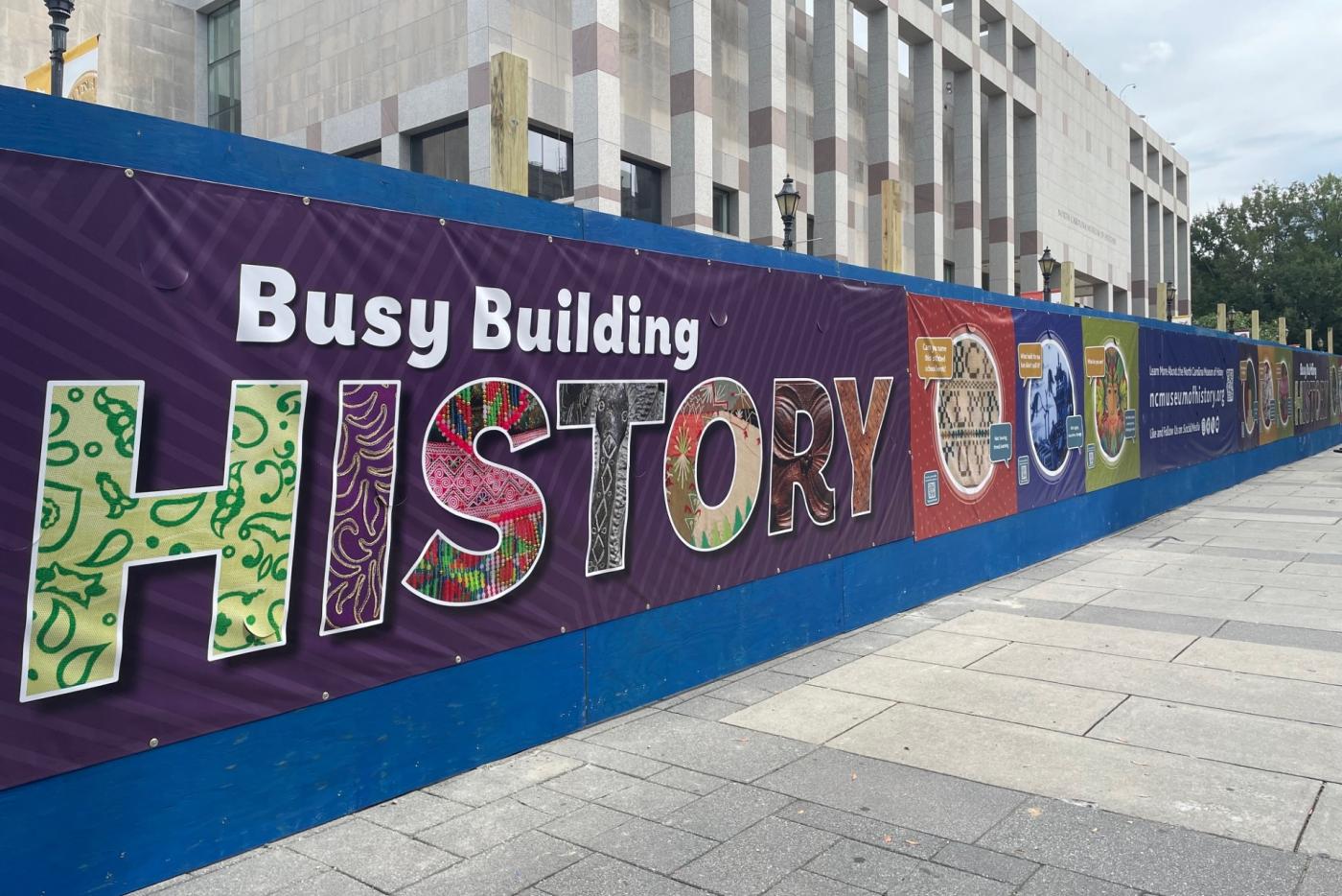 A view of the construction fence outside the North Carolina Museum of History featuring the 'Busy Building History' design.