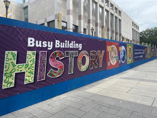 A view of the construction fence outside the North Carolina Museum of History featuring the 'Busy Building History' design.