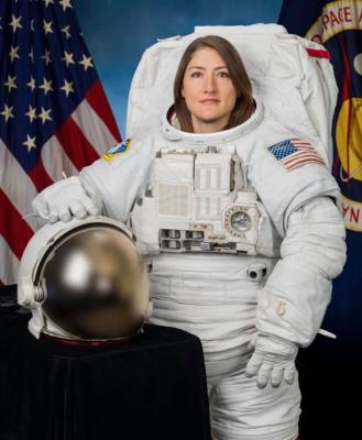 A female astronaut in a NASA spacesuit stands in front of the American flag, with a space helmet placed on a table beside her.