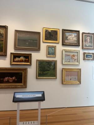 A photo of an exhibit wall at the NCMA with various paintings in gold frames on a white wall. In front of them, there is a touch screen with a sign that reads “Temporarily out of order” on a white stand. There is a silver barrier with a white rope between the wall and the paintings.