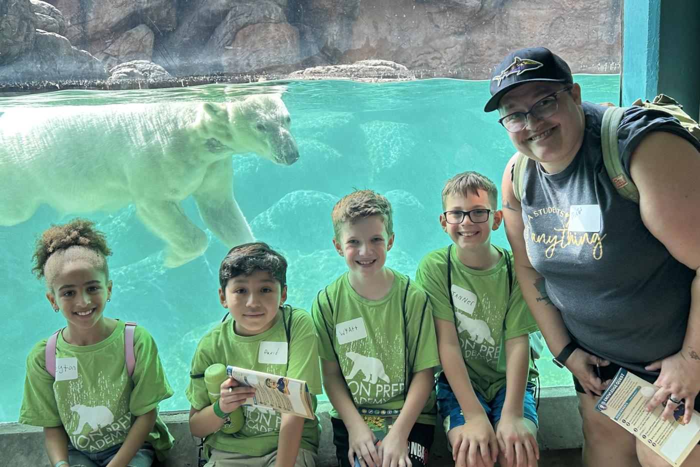Group of children and an adult pose in front of a swimming polar bear at the zoo.
