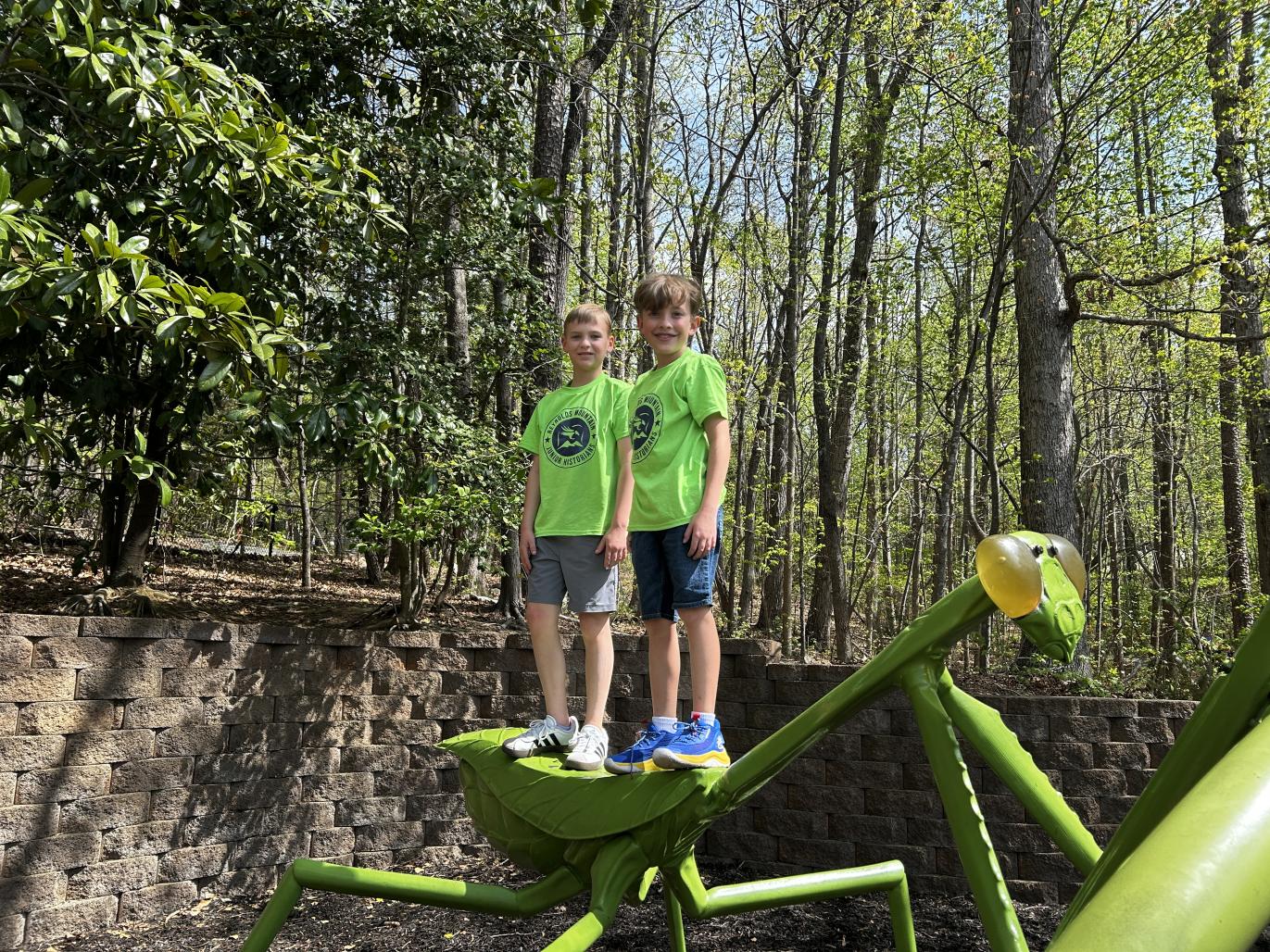 Two children pose with giant insect sculptures at an outdoor exhibit.