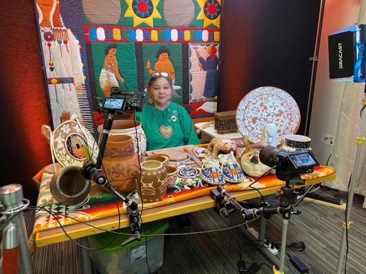 Senora Lynch sits at a table displaying her handcrafted pottery and tools during a virtual education event, with cameras and lighting set up around her.