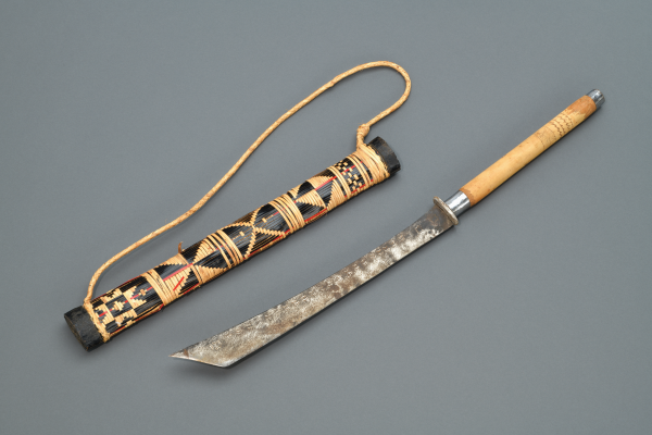 Machete with carved wooden handle and wide blade, shown beside a woven, patterned sheath with carrying cord.