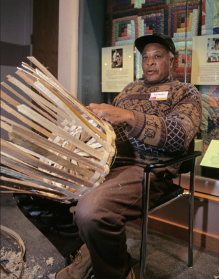 Neal Thomas, wearing a patterned sweater and a black cap, sits in a chair weaving a large basket from wooden strips. 
