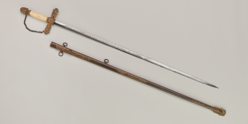 Ornate ceremonial sword with ivory grip, decorative hilt, and engraved blade, shown beside its engraved scabbard.