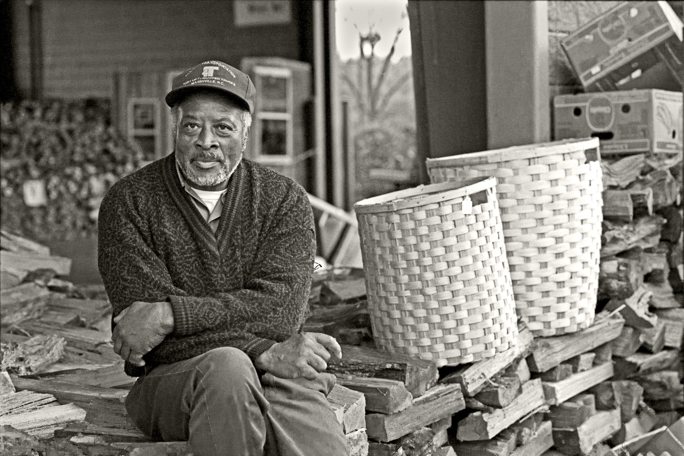 Artist wearing a sweater and a baseball cap, sitting on a pile of firewood with arranged baskets.