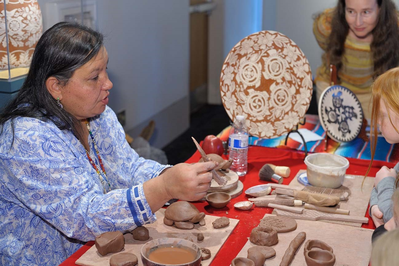 Senora Lynch demonstrates traditional pottery-making at a table with clay, tools, and finished pieces